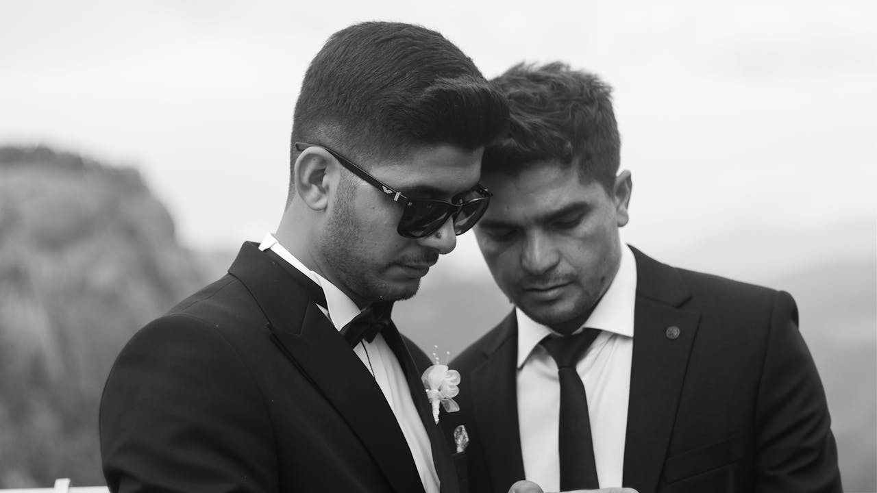 about-01 Two men in suits share a formal moment at an outdoor wedding, depicted in black and white.