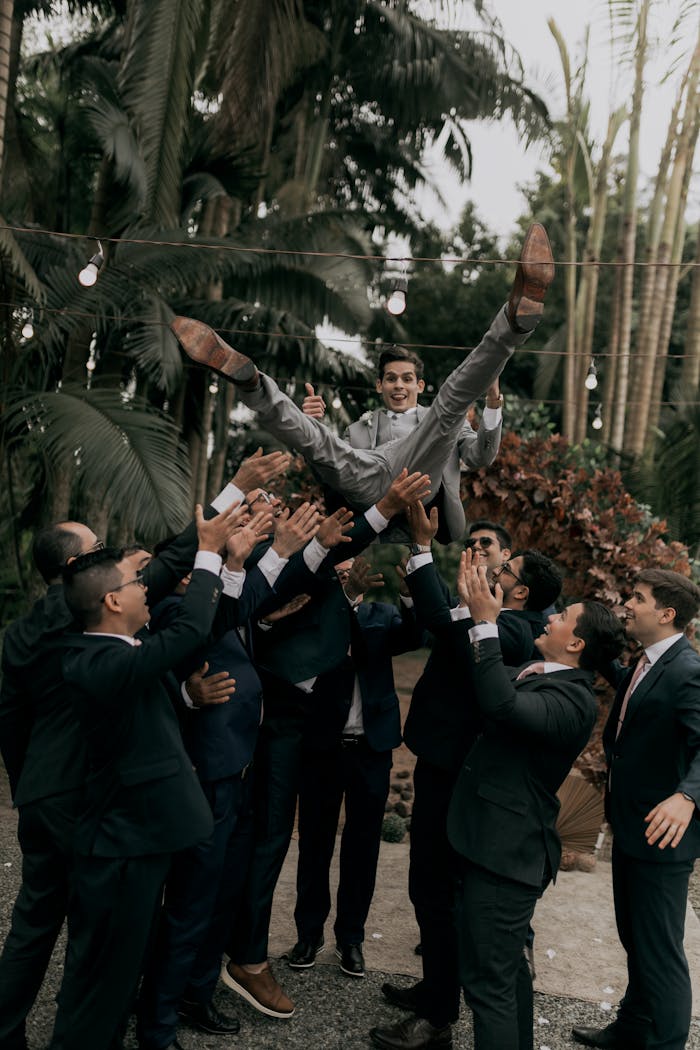 gallery-3 Groomsmen in suits joyfully toss the groom in the air during an outdoor wedding celebration.