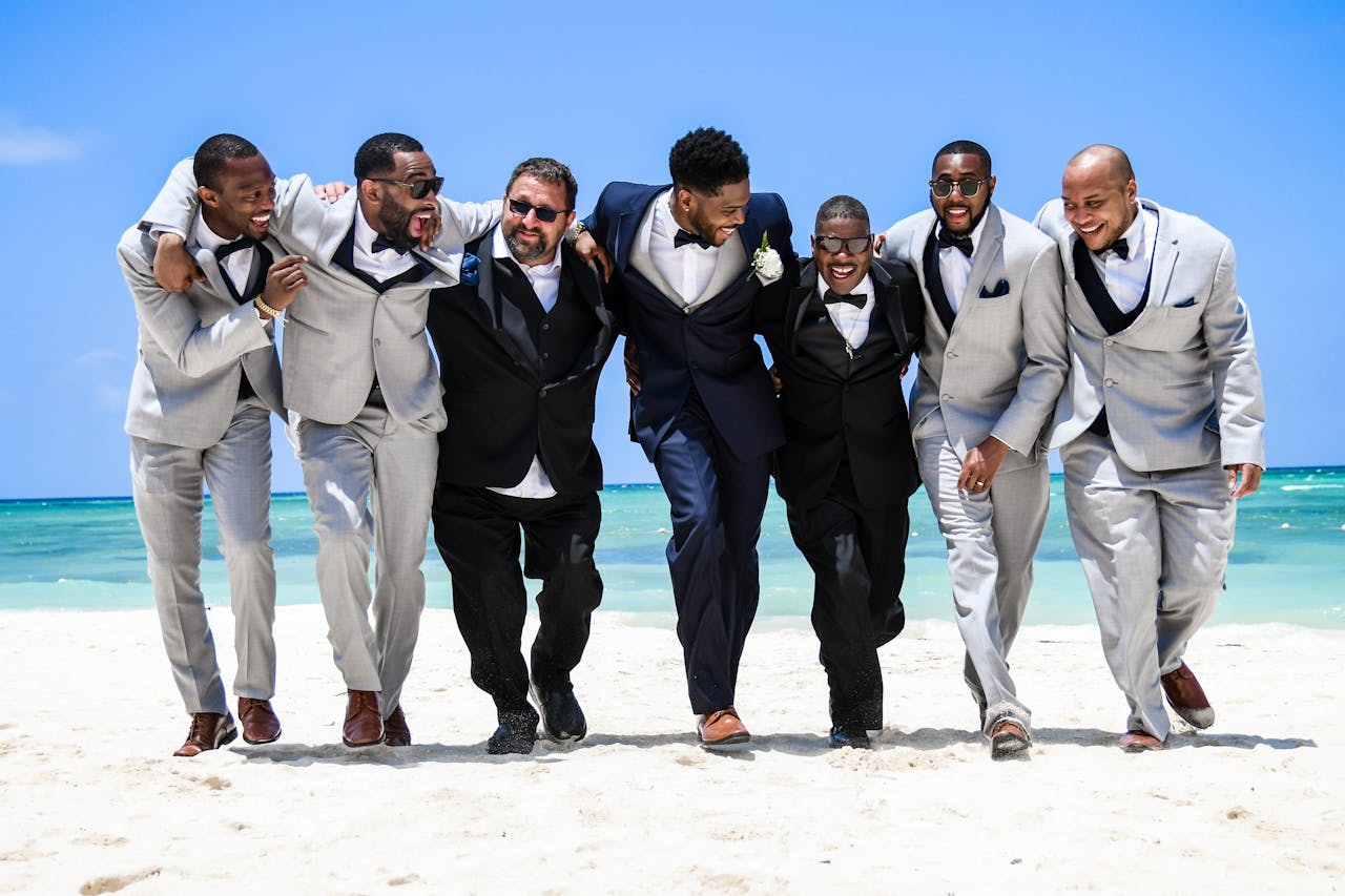 gallery-1 Group of smiling groomsmen enjoying a beach day together, dressed in formal attire.